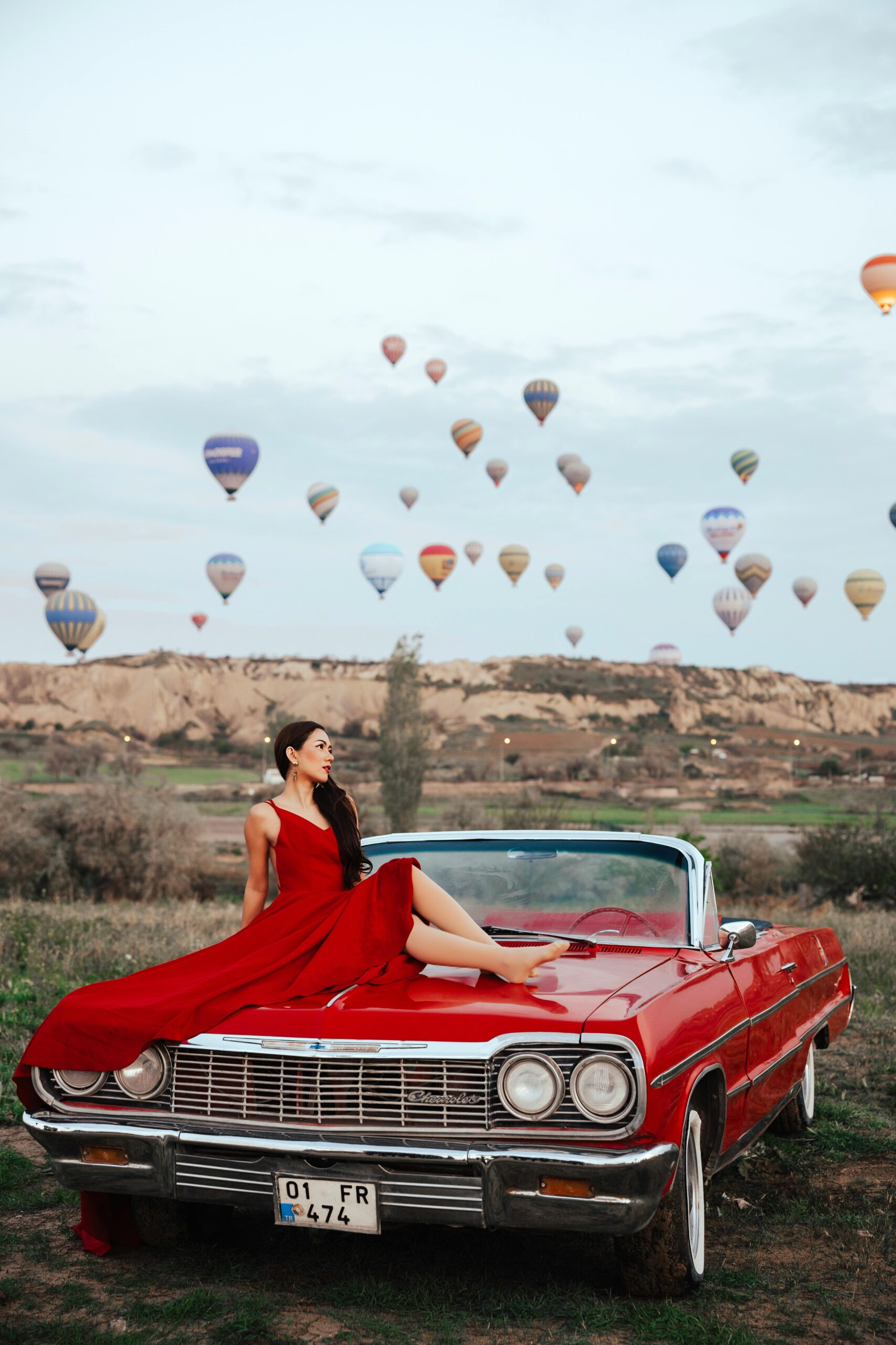 Elegant woman in a red dress poses on a vintage convertible in Cappadocia with hot air balloons in the background.